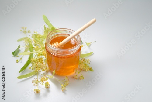 Glass jar filled with natural linden honey decorated with fresh blooming linden flowers and a wooden honey dipper on a white background.