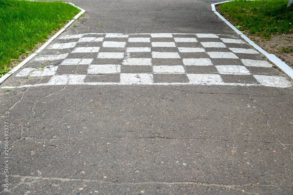 Obraz premium A black and white checkered finish line is painted across a cracked asphalt pathway on an outdoor children traffic training playground.