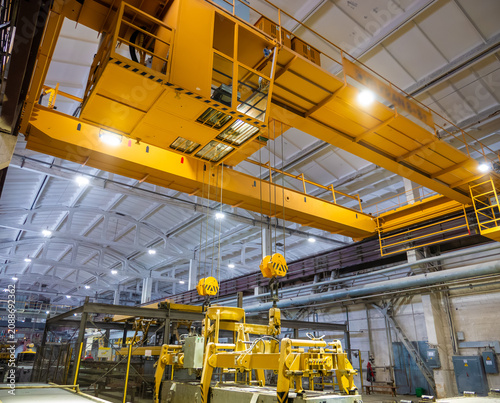 A massive yellow overhead bridge crane with an operator's cabin and heavy-duty lifting hooks spans across the ceiling of a large industrial manufacturing factory workshop.