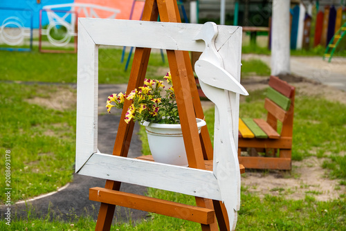 Bild auf Leinwand A creative garden decoration featuring brown wooden artist's easel holding white picture frame with stylized bird silhouette and a pot of blooming yellow and purple petunia flowers on playground lawn