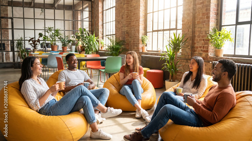 Happy diverse millennial coworkers sit on yellow bean bags in a trendy vibrant coworking space. They hold coffee cups and laugh together, showcasing modern startup culture.