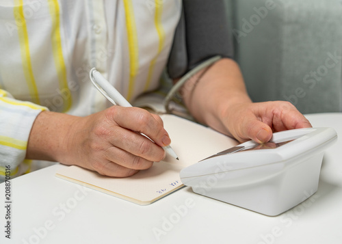 Middle aged woman sitting on sofa at home and writing blood pressure readings in notebook after measurement. Home healthcare, health tracking and preventive care.