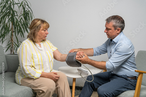 Man measuring blood pressure of middle aged woman sitting on sofa at home. Family care, home healthcare, preventive medicine and cardiovascular health monitoring.