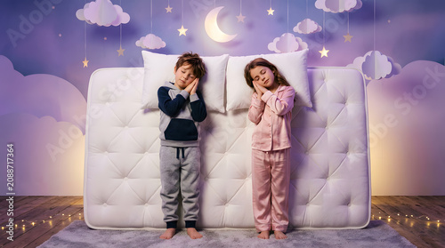 Cute little brother and sister sleeping peacefully on a white mattress with pillows, wearing pajamas against a whimsical starry night background with moon and clouds.