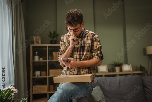 Картината върху платно Young man using smart phone with package anticipating delivery