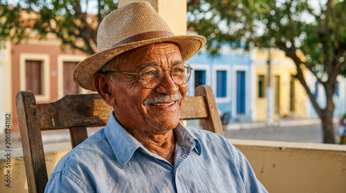 Idoso brasileiro feliz sorrindo, usando chapéu de palha e óculos, sentado ao ar livre em praça colonial colorida