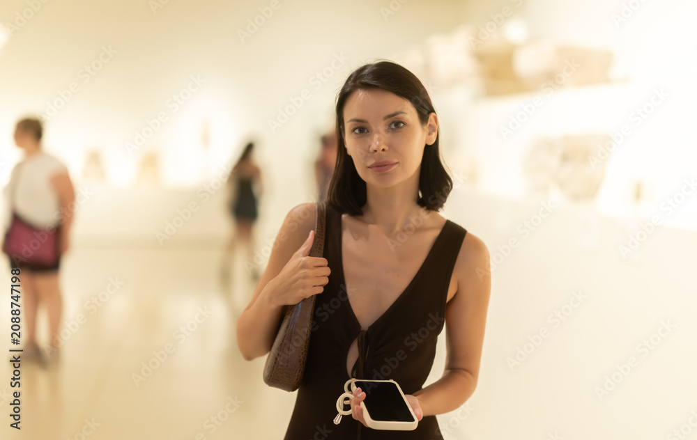 Naklejka premium Girl spends time in museum, examines European antique heritage, hall with columns. Guest of museum walks among exhibits with phone in hands.
