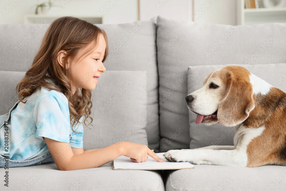 Obraz premium Cute little girl with Beagle dog reading book on sofa in living room