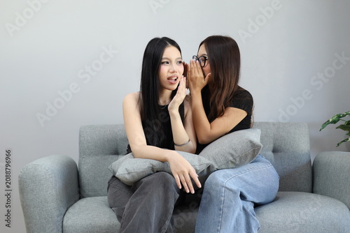 Obraz na plátně A young woman whispers to her friend about shocking news, her friend's reaction is shocked with her mouth open, they are sitting on the sofa at home