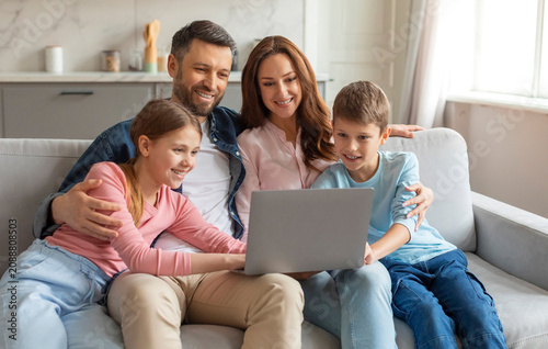 Cheerful young family with kids having fun with laptop at home, watching photos together. Happy parents and their two preteen children using computer while sitting on couch in living room, free space
