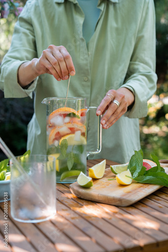 Woman standing in a garden using a long spoon to stir assorted citrus fruits and mint in a glass pitcher of water