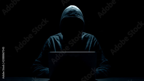Anonymous figure in hooded sweatshirt working on laptop in dark room, dramatic lighting, perfect for cybersecurity, digital crime and tech industry visuals