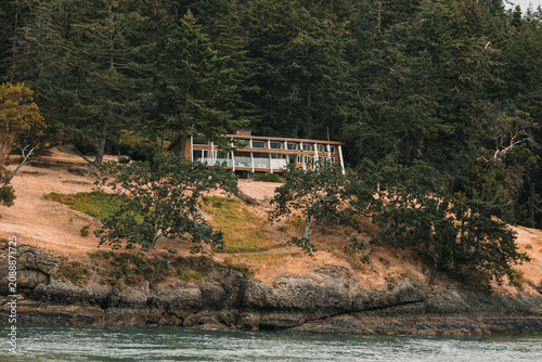 Obraz na plátně A mid-century modern style house situated on a grassy, rocky hillside overlooking the water and surrounded by trees