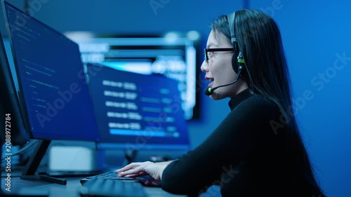 Focused Asian female IT specialist with headset types code on dual monitors in a high tech dark blue server control room. She provides 24 hours, technical support and network cybersecurity monitoring