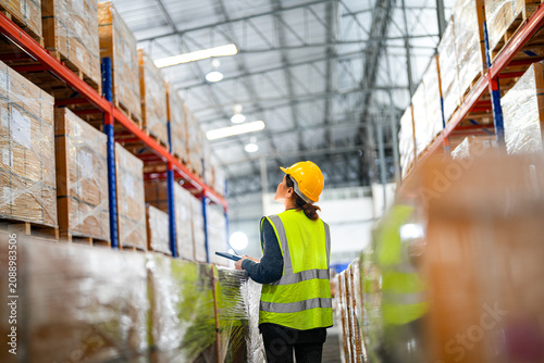 Foto Mature Asian woman working as a warehouse supervisor in protective workwear points at cartons while auditing the stock in a large fulfilment centre with rows of stacked pallets on an