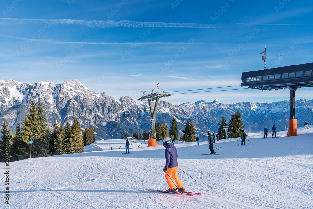Fototapeta premium Skiers enjoy a sunny day on the groomed slopes near the modern ski lift station in Saalbach, Austria. The majestic snowy Alps provide a stunning backdrop for winter sports.
