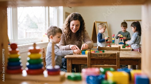 A smiling female teacher assists a young boy with colorful wooden blocks in a bright preschool classroom, with other children playing in the background.