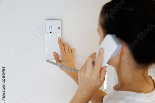 Konstfotografi Woman pressing the button on a wall mounted home intercom while talking through the handset