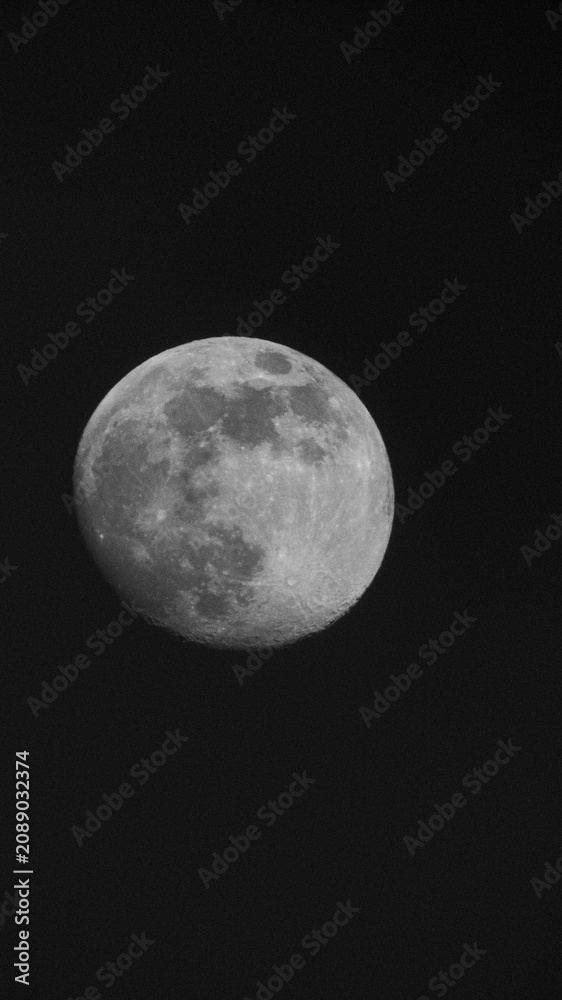 Fototapeta premium This image features a stunning close-up shot of the moon in its gibbous phase, nearing a full moon. The entire sunlit surface of the moon beams brilliantly against a pitch-black, starless night sky. 