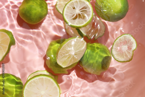Canvas Print Top view of fresh whole green limes and slices floating in clear rippling water with a pink background