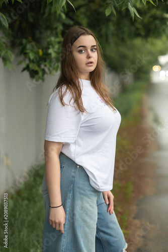 Full-length portrait of a confident young plus-size woman posing on a wet asphalt road. She wears a casual white t-shirt and trendy baggy blue ripped jeans, looking directly into the camera.