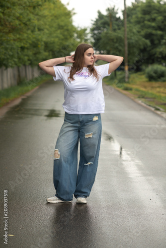 Full-length portrait of a confident young plus-size woman posing on a wet asphalt road. She wears a casual white t-shirt and trendy baggy blue ripped jeans, looking directly into the camera.