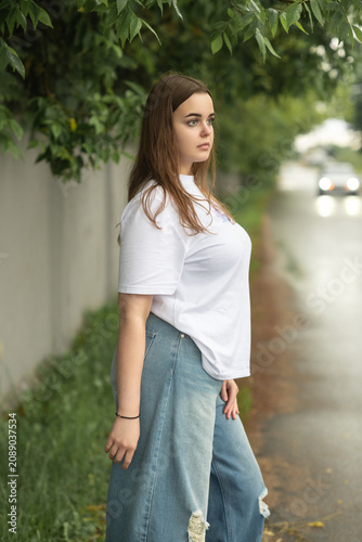 Full-length portrait of a confident young plus-size woman posing on a wet asphalt road. She wears a casual white t-shirt and trendy baggy blue ripped jeans, looking directly into the camera.