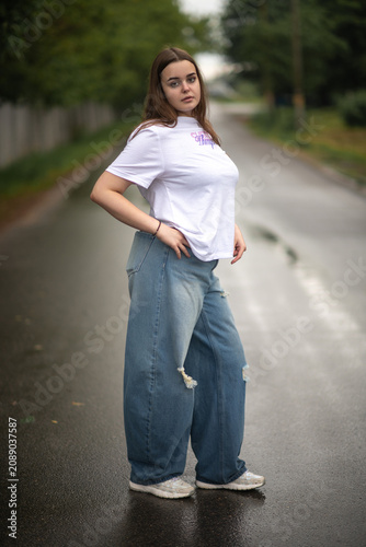 Full-length portrait of a confident young plus-size woman posing on a wet asphalt road. She wears a casual white t-shirt and trendy baggy blue ripped jeans, looking directly into the camera.