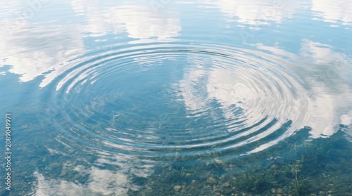 Circular ripples on clear mountain lake with submerged rocks and sky reflections