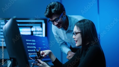 Two tech asian engineers collaborate on an innovative robotics project inside a dark futuristic high-tech lab. A female developer holds a small robot and discusses the data with a male colleague.