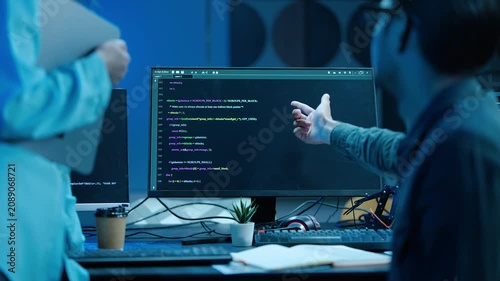 Two software developers collaborating on a coding project late at night in a dark modern office. A male programmer points at the computer screen, explaining complex algorithms to a colleague.