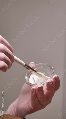 Professional cosmetologist hands in pink gloves mixing cosmetic chemical peel or white enzyme cream mask with a brush in a bowl before treatment for a client. Vertical shot. Close-up