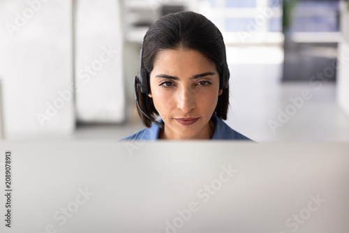 Closeup of female support agent in headset assisting client remotely  looks at computer monitor  listens attentively  providing professional customer or technical support services. Helpdesk workflow