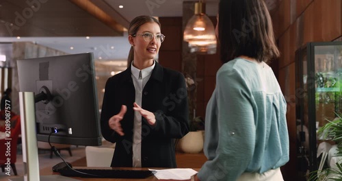 Pretty woman receptionist dressed in formal attire stands at reception front desk and serves female client, making check-in, explain hotel check-out rules, provide helpful information. Guests services