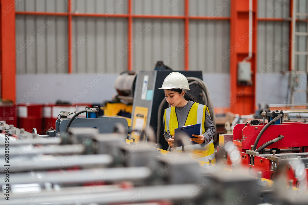 Fototapeta premium Female quality engineer inspecting industrial production equipment while recording operational data on clipboard inside modern factory. Manufacturing inspection supporting quality assurance.