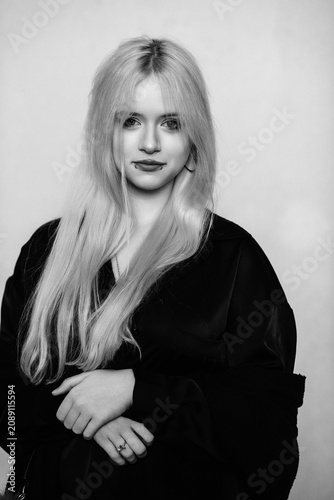 Black and white portrait of a young blonde woman with alternative style, lip piercing and a skull necklace looking away against a plain background.
