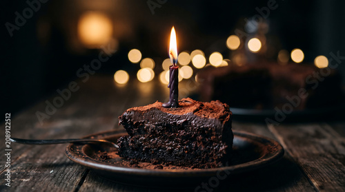 Dark decadent chocolate cake slice with burning candle on rustic