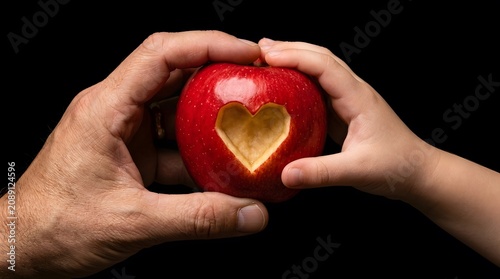 Generational connection concept featuring a red apple with a carved heart held by adult and child hands against black