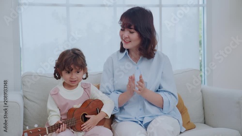 A mother sits on a couch with her young daughter. The girl plays a ukulele while her mom looks at her and shares a smile. They enjoy a happy moment together in their home.
