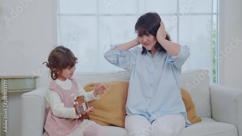 A mother sits on a couch with her young daughter. The girl plays a ukulele while her mom looks at her and shares a smile. They enjoy a happy moment together in their home.