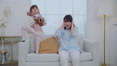 A mother sits on a couch with her young daughter. The girl plays a ukulele while her mom looks at her and shares a smile. They enjoy a happy moment together in their home.