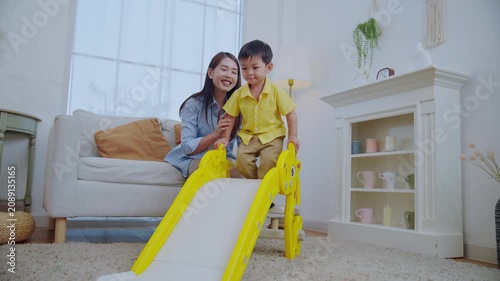 A mother sits beside her young son as he plays on a small slide. They are in a bright living room with a sofa and plants. They share smiles and laughter together.