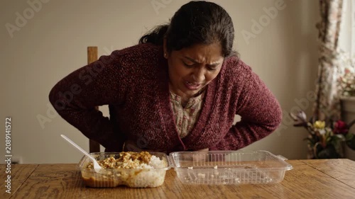 Woman clutching stomach pain sitting wooden table with food container and spoon showing discomfort distress possible indigestion or food poisoning