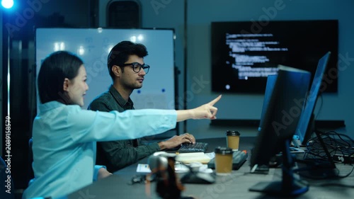 Diverse tech professionals collaborate on complex software development in a dark office. Asian female programmer points at a computer screen with code while a male developer types late at night.