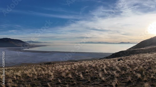 Scenic footage of Antelope Island and Great Salt Lake in Utah during sunset. The video features a smooth camera pan to the left, showing a grassy hill slope and mountains in a hazy golden hour light.