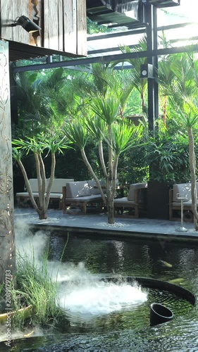 White mist rising from circular pond beside tropical lounge with palm trees, cushioned seating, pergola beams, concrete deck, sunlit reflections and calm resort garden mood