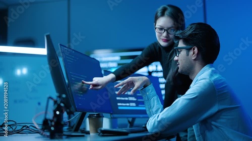 Two asian IT professionals analyze complex data on multiple computer screens in dark, high-tech server room. Diverse male and female team discuss cybersecurity code and business intelligence software