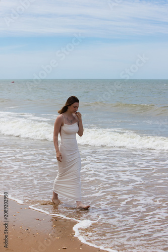 Obraz na płótnie A young, beautiful woman in a long white dress walking along the beach on a windy day