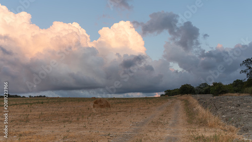 Wide field with hay bales and glowing clouds above a rural dirt road.