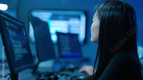 Focused young Asian woman software engineer working on computer code in a dark IT office with blue lighting. Professional female programmer analyzing network data and developing digital security.